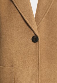 Abrigo de mezcla de lana marrón que presenta un cuello con muesca, un cierre con un solo botón negro y un bolsillo lateral. Acabado suave y texturizado.