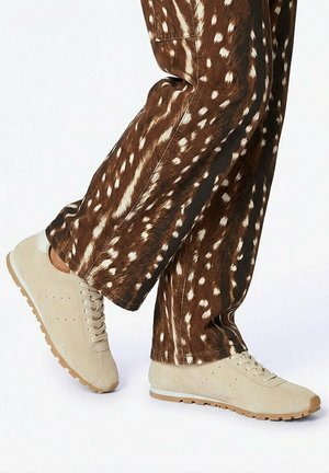 Personne portant des baskets en daim beige et un pantalon marron à imprimé animalier blanc tacheté, debout avec une jambe croisée sur l'autre.