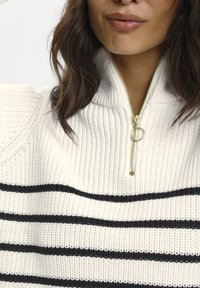 Suéter de punto blanco con rayas horizontales negras, con un cuello con cremallera y una textura acanalada. Detalles en dorado en la cremallera.