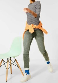 Randig långärmad skjorta, orange tröja knuten i midjan, gröna texturerade byxor, blå strumpor och vita sneakers. En mintgrön stol i närheten.