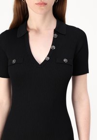 Robe courte noire à manches courtes côtelées avec un col en V, ornée de boutons décoratifs de couleur argentée et deux poches près de la poitrine.