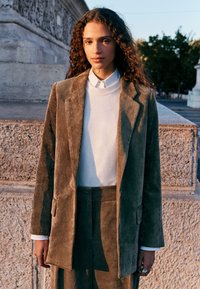 Brown corduroy blazer with textured vertical stripes, paired with a white sweater and matching pants, featuring a relaxed fit and pockets.