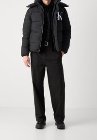 Doudoune noire matelassée avec capuche et fermeture éclair, arborant un logo blanc sur la poitrine. Associée à un pantalon noir et des baskets.