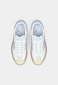 Baltas Adidas kedas ar gaiši ziliem svītrām, krēmkrāsas purngaliem, brūniem zoleiem un ar zīmolu apdrukātām iekšpusēm, skatīti no augšpuses uz baltas fona.