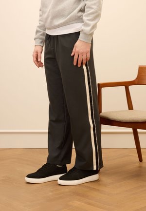 Pantaloni da allenamento grigio scuro con strisce bianche sui lati, realizzati in tessuto morbido. Abbinati a sneakers nere; il modello si trova accanto a una sedia di legno.