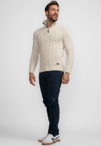 Cremfarbener Kabelstrickpullover mit Reißverschlusskragen und Holztoggle-Detail, kombiniert mit navyblauen Hosen und weißen Sneakers mit schwarzen Akzenten.