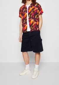 Monivärinen lyhythihainen paita, jossa on abstrakteja kuvioita oranssina, vaaleanpunaisena ja mustana, yhdistettynä tummansinisiin vakosamettishortseihin ja valkoisiin lenkkareihin.