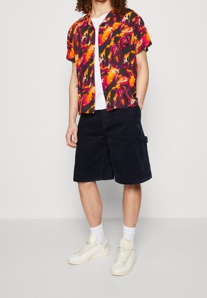 Bunt gestreifte kurzärmelige Hemd mit abstrakten Mustern in Orange, Pink und Schwarz, kombiniert mit marineblauen Cordshorts und weißen Turnschuhen.