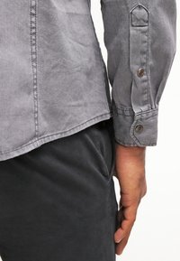 Grijze shirt met een shirtstaartzoom, knoopmanchetten en een gestructureerde stof. Gecombineerd met donkere broeken, die een slanke pasvorm tonen.