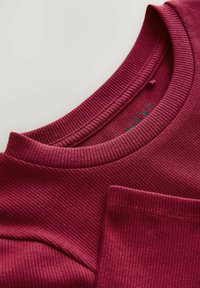 Pull en ribbed bordeaux avec un col rond et une poche sur la poitrine. Texture lisse avec un léger éclat, mettant en valeur des coutures soignées.