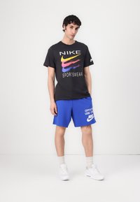 Tricou din bumbac negru cu logo Nike multicolor, asortat cu șorturi albastre ce prezintă un imprimeu alb cu text Nike. Adidașii albi completează ținuta.