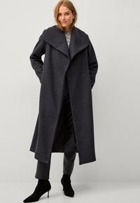 Lungo cappotto di lana grigio scuro con colletto ampio, davanti doppiopetto e taglio dritto. Indossato con pantaloni in pelle nera e stivali a punta.