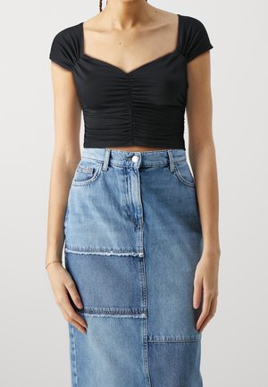 Woman wearing a ruched black crop top with cap sleeves and a high-waisted patchwork denim skirt against a plain background.