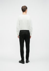 Muž stojaci chrbtom, oblečený v bielej košeli s dlhým rukávom, čiernych nohaviciach a čiernych topánkach, pred jednoduchým svetlým pozadím.