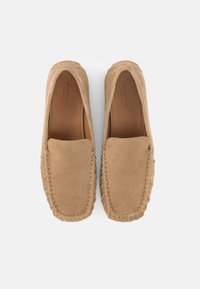 Μπεζ σουέτ loafers τύπου slip-on με ραφές διακόσμησης, επενδύσεις εσωτερικά με λογότυπο και στρογγυλεμένο σχήμα στα δάχτυλα, τοποθετημένα παράλληλα σε λευκό φόντο.