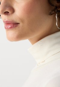 Cream high-collared top made of soft fabric. The collar features an embroidered logo detail. Smooth texture with a fitted design.