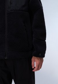 Black fleece jacket with textured, shaggy fabric. Features a zippered pocket and a smooth upper section. Includes brand logo on the sleeve.