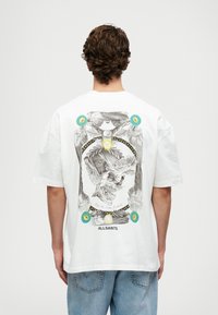 Λευκό βαμβακερό t-shirt με σχέδιο γραφικών που απεικονίζει έναν κρανίο με φτερά, περιτριγυρισμένο από πολύχρωμα στοιχεία και κείμενο. Χαλαρή γραμμή με κοντά μανίκια.