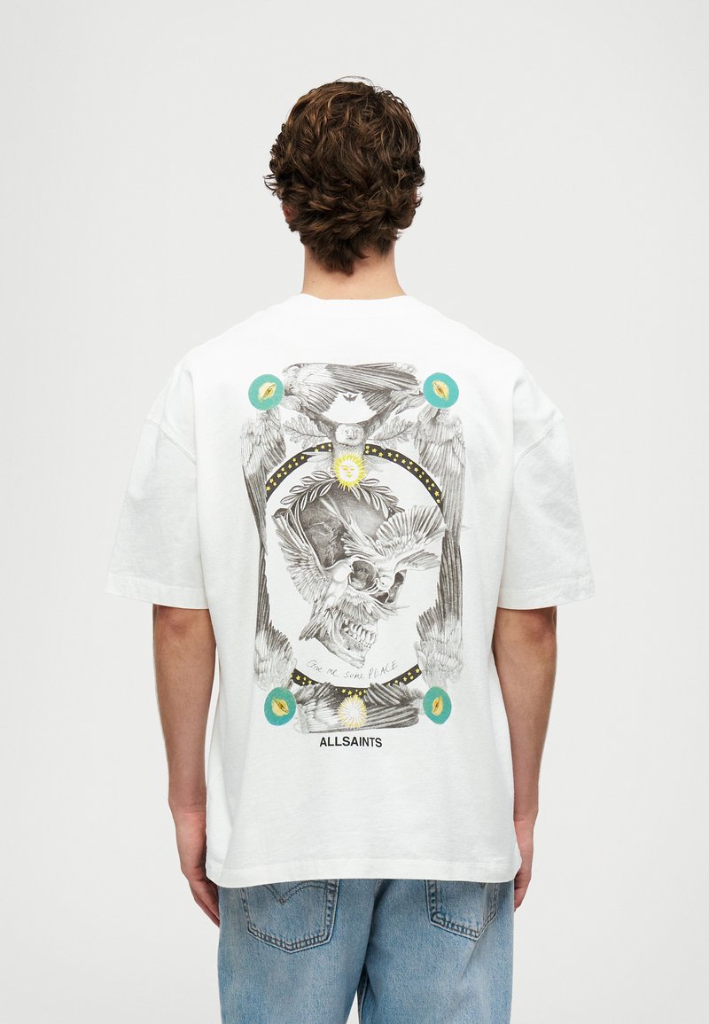 Λευκό βαμβακερό t-shirt με σχέδιο γραφικών που απεικονίζει έναν κρανίο με φτερά, περιτριγυρισμένο από πολύχρωμα στοιχεία και κείμενο. Χαλαρή γραμμή με κοντά μανίκια.