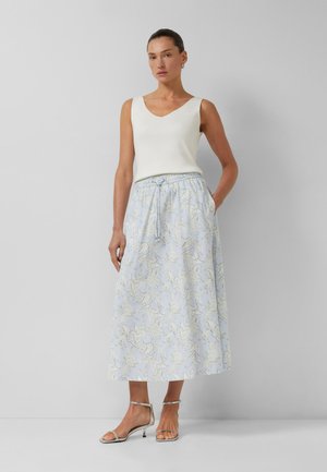 Kobieta stoi, ubrana w białą bluzkę bez rękawów, niebieską kwiecistą sukienkę midi z sznurowanym paskiem w talii oraz srebrne sandały na obcasie z paskami, na jednolitym szarym tle.