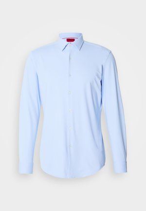 Lys blå skjorte med lange ærmer lavet af glat stof, med knaplukning foran, spredt krave og rundet kant. Mærketiketten er synlig indeni kraven.