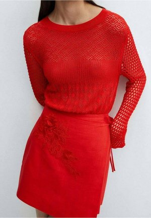 Kobieta ubrana w czerwoną, wzorowaną sweter dziany z motywami diamentów i siatki, zestawiony z czerwoną spódnicą ozdobioną kwiatową haftem po boku.