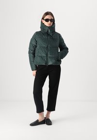 Groene, pufferjack met hoge kraag en ritsluiting. Gelaagd oppervlak en vierkante vorm. Model draagt zwarte broeken en elegante schoenen.