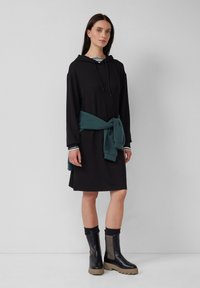 Schwarzes Kapuzenkleid mit langen Ärmeln, einer verstellbaren Kapuze und lockerer Passform. Ein türkisfarbenes Sweatshirt, das um die Taille gebunden ist, und schwarze Knöchelstiefel mit strukturierten Akzenten.