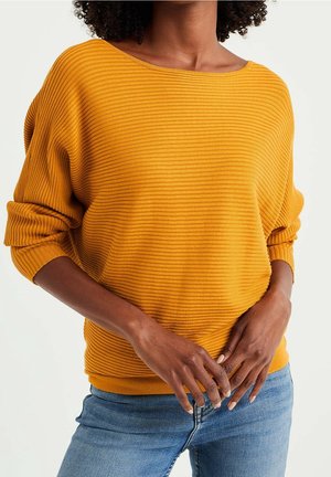 Frau trägt einen senfgelben gerippten Langarmpullover mit hochgekrempelten Ärmeln und blaue Jeans, die Hände locker vor dem Körper verschränkt.