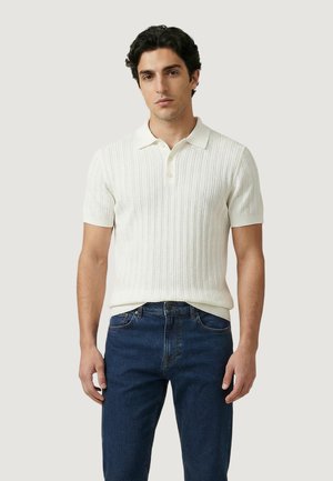 Homme debout portant un polo blanc à manches courtes côtelé et un jean bleu foncé, sur un fond uni clair.