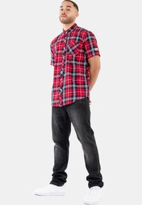 Košile s krátkým rukávem v červené a černé kostce s knoflíkovým zapínáním, spárovaná s tmavými džínami a bílými teniskami. Výrazný detail kapsy na levém prsu.
