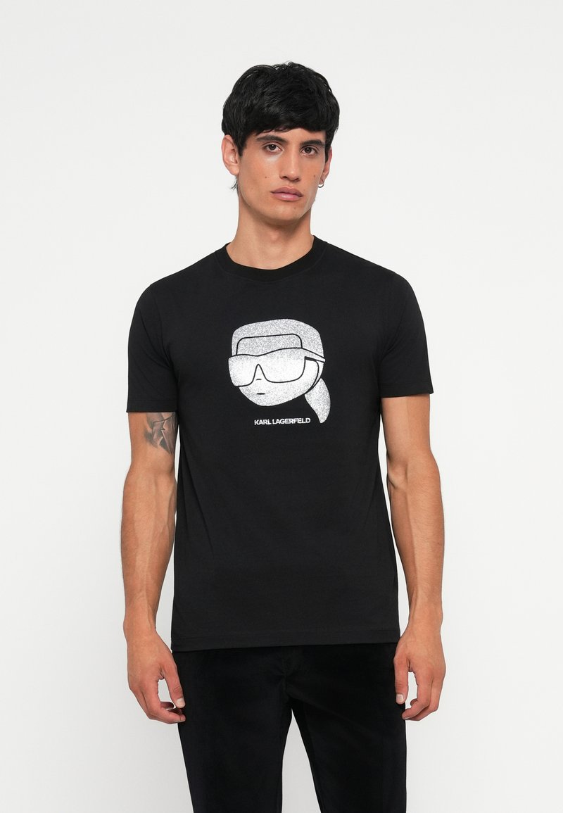 Čierne bavlnené tričko s striebornou grafikou postavy v nadmerných slnečných okuliaroch s textom "KARL LAGERFELD" pod ním.