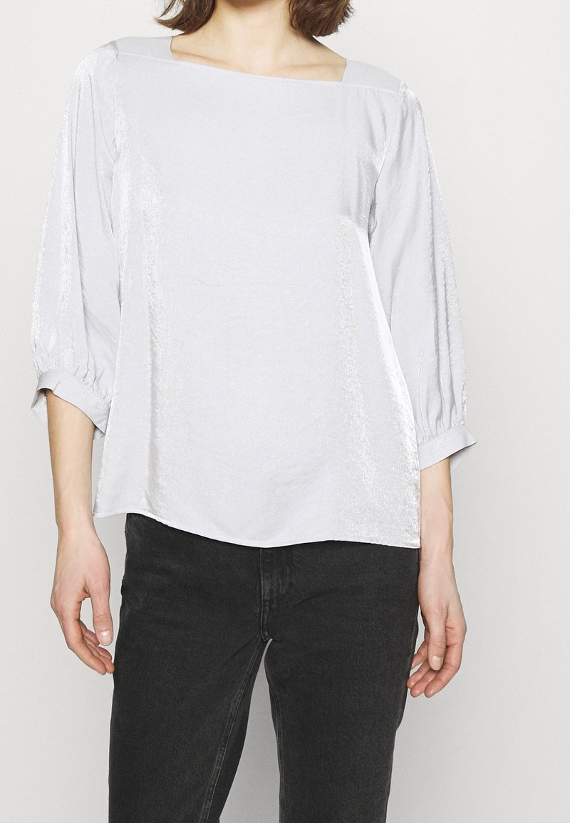 Femme portant un chemisier blanc ample avec des manches trois-quarts froncées et un pantalon noir taille haute sur un fond uni.