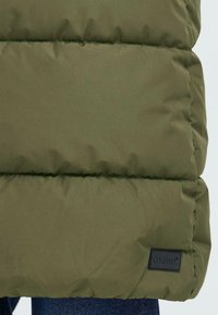 Olivgrüne, gepolsterte Jacke mit gestepptem Muster, ausgestattet mit einem kleinen schwarzen Markenlabel in der unteren rechten Ecke. Jeansstoff ist darunter sichtbar.