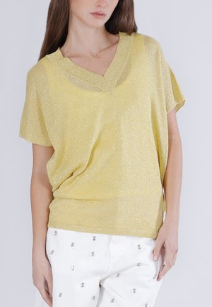 Femme portant un haut jaune ample et scintillant à col en V et un pantalon blanc orné de petites clous argentés, debout devant un fond uni.