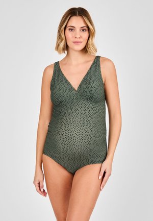 Femme enceinte portant un maillot de bain de maternité sans manches vert à pois noirs, debout devant un fond gris clair uni.