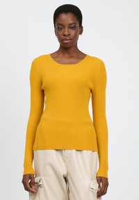 Maglione giallo a coste con maniche lunghe e scollo a barca, abbinato a pantaloni chiari plissettati.
