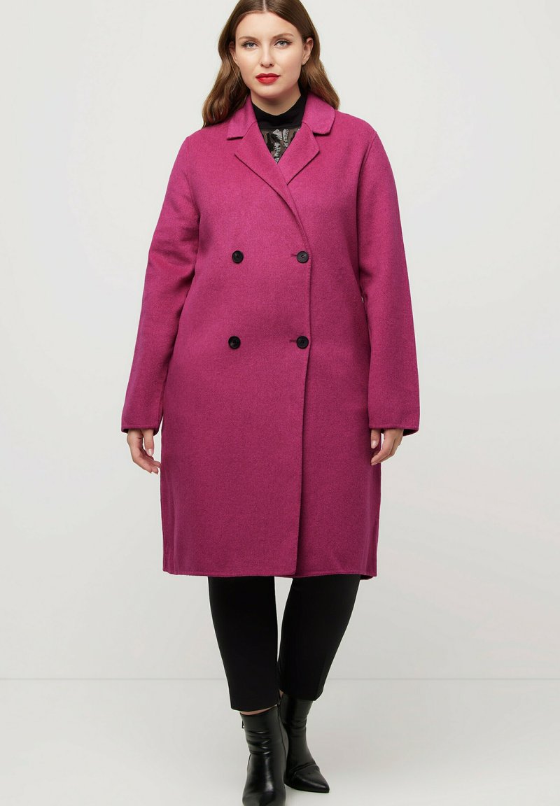 Cappotto doppiopetto in misto lana fucsia con colletto a ribalta, bottoni neri e silhouette dritta. Abbinato a pantaloni neri e stivali.