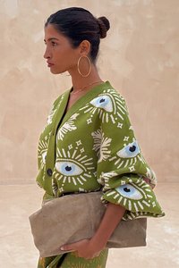 Cardigã de malha verde com padrões de olhos, mangas oversized e decote em V. Acessório é uma carteira bege. Textura subtil e cores contrastantes.