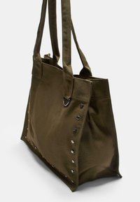 Olivgrüner Canvas-Shopper mit zwei Schulterriemen, verziert mit Metallnieten und Sternakzenten auf einer Seite. Rechteckige Form, strapazierfähige Textur.