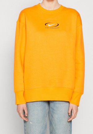 Osoba ubrana w jaskrawo pomarańczową bluzę Nike Sportswear z czarnym logo na piersi oraz jasnoniebieskie podarte dżinsy, stojąca na tle jednolitego tła.