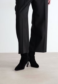 Zwarte hoge hak enkellaarsjes van zacht suède met een spitse neus en slanke stilettohak, gecombineerd met op maat gemaakte zwarte broeken.