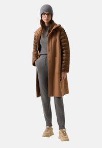 Brauner langer Mantel mit steppähnlichen Ärmeln, grauer Pullover und passende Jogginghose. Graue Strickmütze und beigefarbene Sneakers runden das Outfit ab.