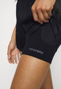 Czarne sportowe szorty mają dopasowaną podszewkę, logo na udzie oraz teksturowaną tkaninę z widocznym przeszyciem i detalem bocznej kieszeni.