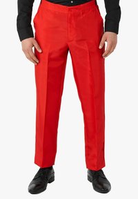 Pantalones rojos ajustados con una textura suave, pinzas delanteras y cierre de botón, combinados con zapatos negros y una camisa negra de manga larga.