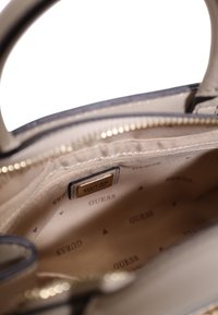 Interno di una borsa color beige chiaro con texture in satin, dotato di una cerniera, hardware dorato e un'etichetta con logo. Motivo di branding sottile sulla fodera.