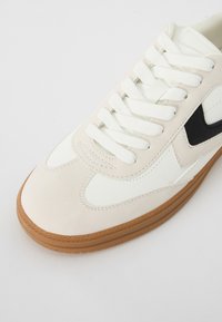 Biało-beżowy sneaker z gładką skórzaną cholewką, teksturowanymi zamszowymi akcentami, czarnym logo oraz okrągłą brązową gumową podeszwą.