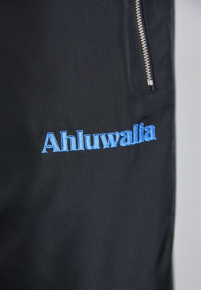 Crna tkanina s izvezenim plavim tekstom "Ahluwalia." Ima srebrni patent zatvarač sa strane. Glatka tekstura s minimalističkim dizajnom.