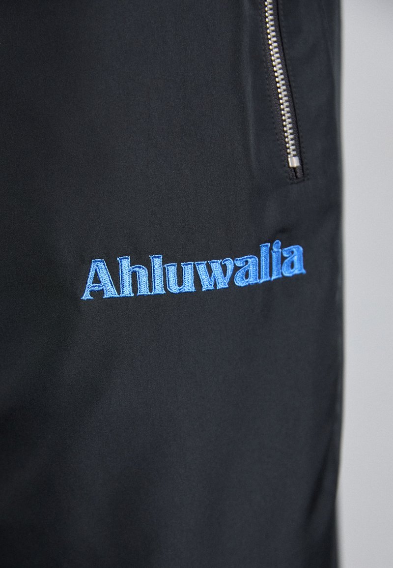Tissu noir avec le texte brodé en bleu "Ahluwalia." Présente une fermeture éclair argentée sur le côté. Texture lisse avec un design minimaliste.