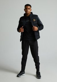 Giacca imbottita nera con colletto alto, chiusura con zip e logo sul petto, abbinata a una felpa nera e pantaloni coordinati, indossata con sneakers nere.